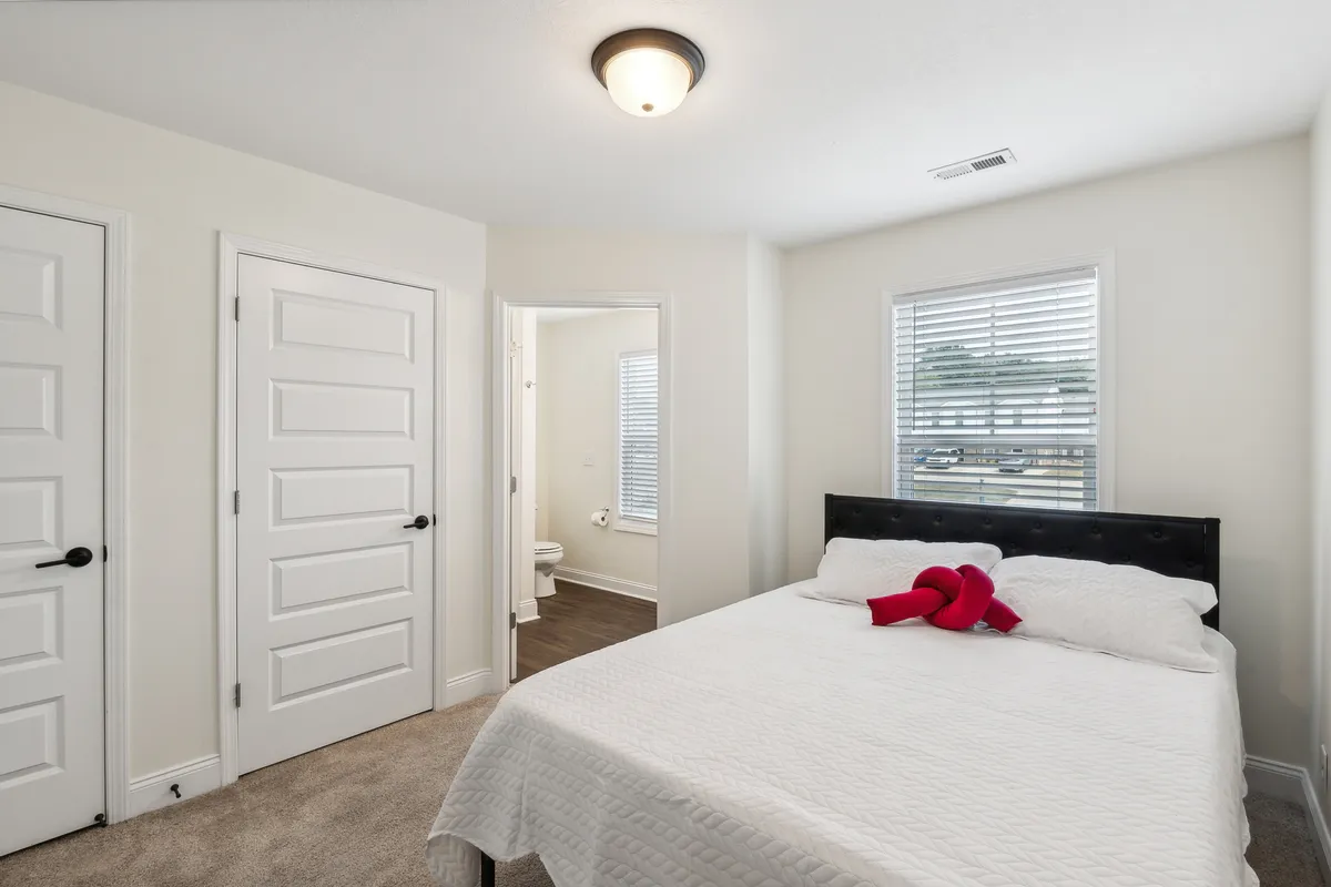 A tranquil bedroom with a neatly made bed adorned with decorative pillows, ample windows with blinds, and doors leading to other rooms. The space is uncluttered, filled with natural light, and exudes calmness and privacy.