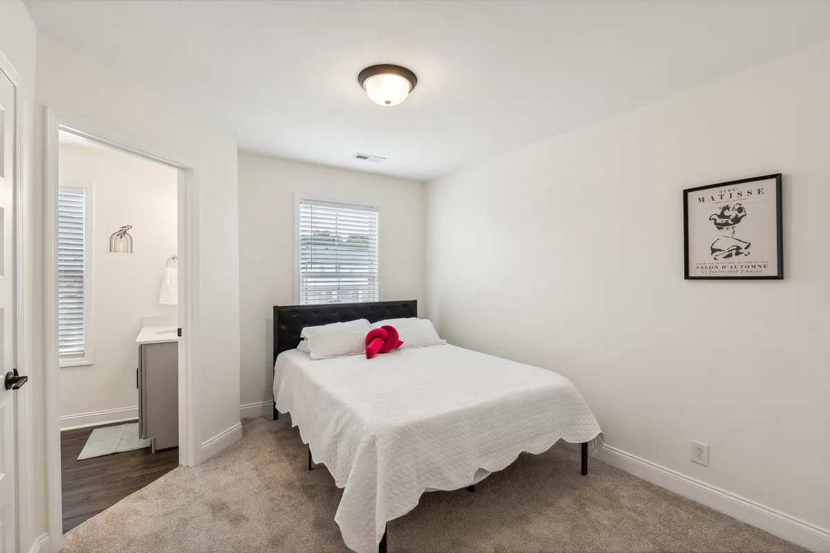 A serene bedroom bathed in natural light, featuring a cozy bed with crisp white bedding and a bold red accent pillow. The large window provides a sense of openness and connection to the outdoors, making the space feel bright and inviting.