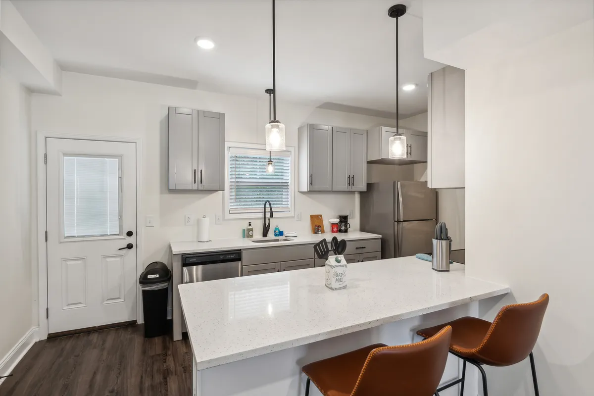 This image showcases a modern, inviting kitchen featuring a kitchen island with bar stools, a sink, window, and visible appliances. The scene exudes a welcoming atmosphere, suggesting a social space where cooking, entertaining, and casual dining occur.