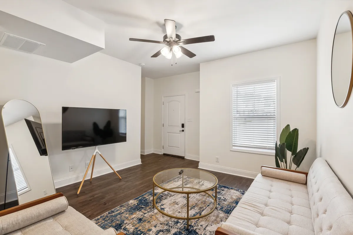 The contemporary living room features a plush sofa, coffee table, television, and decorative touches like a mirror and plant, all arranged for relaxation and socializing. The layout suggests a welcoming space where guests can immediately unwind upon arrival.