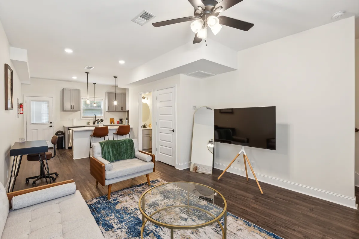 This image presents a cozy living area with a sofa, coffee table, TV, and bar stools at a kitchen island in the background. The space is open-plan, seamlessly connecting the living and kitchen zones, and is styled in a contemporary, inviting way ideal for relaxing or socializing.