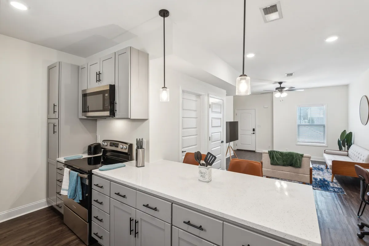 A sleek, contemporary kitchen with a central island leads directly into the adjoining living area, creating a bright, connected space. Stainless steel appliances, modern cabinetry, and bar stools suggest both convenience and style for gatherings or family meals.