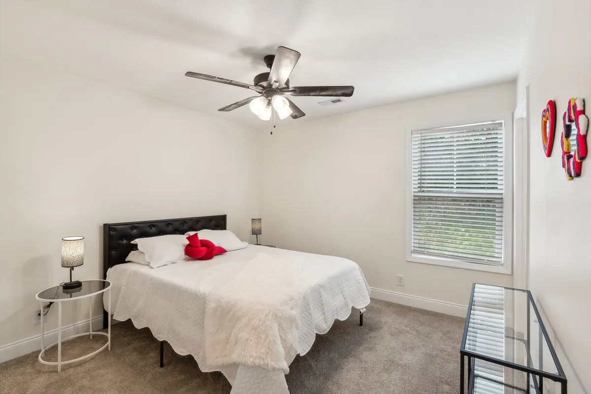 A cozy bedroom featuring a bed with crisp white bedding and decorative red heart pillows, complemented by a glass side table with a lamp, and a window with blinds letting in soft natural light. The décor is soothing and the layout suggests ample space for relaxation and restful nights.