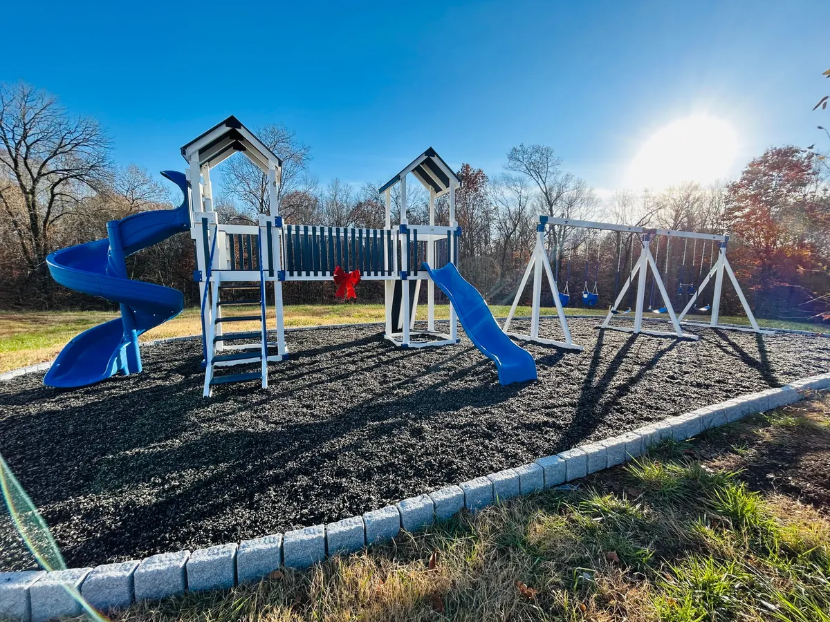 This image showcases a lively playground scene with a blue slide, swing set, and playful red decorations. The structure invites children to climb, slide, and swing, while the open outdoor space suggests room for family gatherings and active play.
